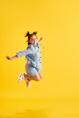 Full-length portrait of active little kid, girl, child in casual clothes cheerfully jumping against yellow studio background. Concept of emotions, childhood, education, fashion, lifestyle, ad
