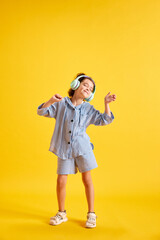 Full-length portrait of beautiful little child, girl listening to music in headphones against yellow studio background. Concept of emotions, childhood, education, fashion, lifestyle, ad