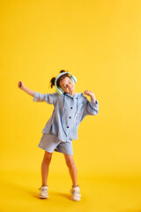 Fototapeta na wymiar Full-length portrait of beautiful little child, girl listening to music in headphones and dancing against yellow studio background. Concept of emotions, childhood, education, fashion, lifestyle, ad