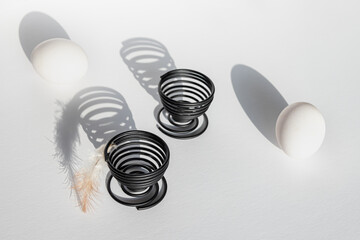 two white eggs lie next to spiral-shaped metal black stands casting shadows on a white background,