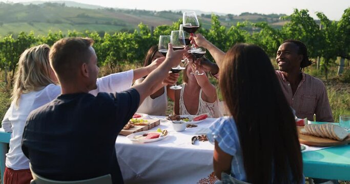 Happy adult friends having fun cheering with red wine and eating together outdoor - Vineyard in background