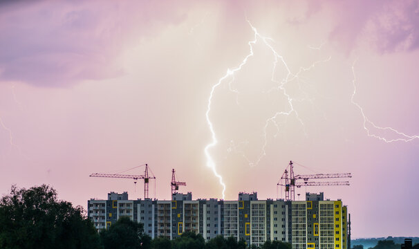 An apartment building with construction cranes against the background of the night sky where you can see lightning
