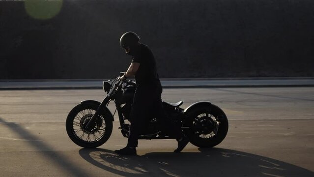 Silhouette of a man with a motorcycle moving on the asphalt. A biker in a black T-shirt and helmet carries a bike in his hands. The man's bike broke. Sad biker walks with a broken motorcycle.