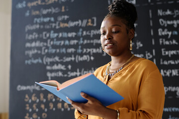African American teacher reading book at IT lesson standing against blackboard
