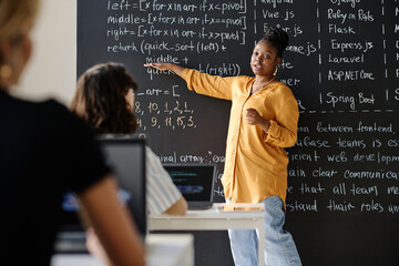 African American teacher pointing at blackboard with IT formulas and explaining material to...