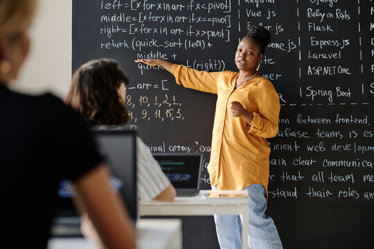 African American teacher pointing at blackboard with IT formulas and explaining material to students at class