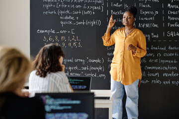 Teacher standing against the blackboard and teaching IT lesson to students at class