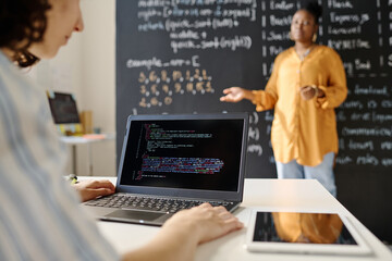Rear view of student sitting at desk and typing codes on laptop during IT lesson