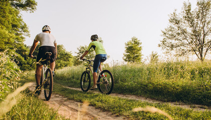 A man and a woman ride bicycles on a dirt road in the summer at sunset. Active rest in the summer on bicycles. - obrazy, fototapety, plakaty