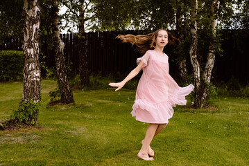 A little girl with long brown hair in a long pink dress dances and jumps on a well-trimmed lawn in the garden of a small house in the village in summer