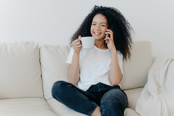 Cheerful Afro American on living room couch, coffee break, calls friend with modern gadget, happy smile, engaging conversation. Tech-lifestyle blend.