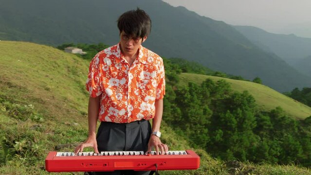 Static shot of young man playing the piano surrounded by hilly terrain covered with lush green vegetation at daytime.