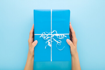 Young adult woman hands holding big wrapped gift box with white ribbon on light blue table...