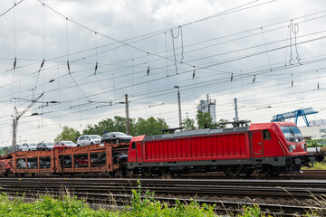 Red locomotive pulling railway wagon with autocars.. The sky at the intersection of wires is covered with clouds.