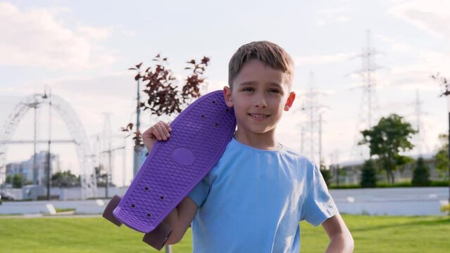 A happy boy is relaxing in the park riding a skateboard. The child looks at the camera, smiles and holds a skate board on his shoulder. Active recreation of children, sports. Kid dreams.