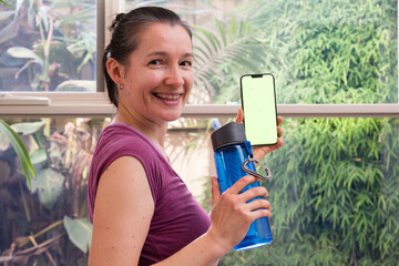 Portrait of smiling woman in sport clothing with bottle of water showing her phone with green screen.