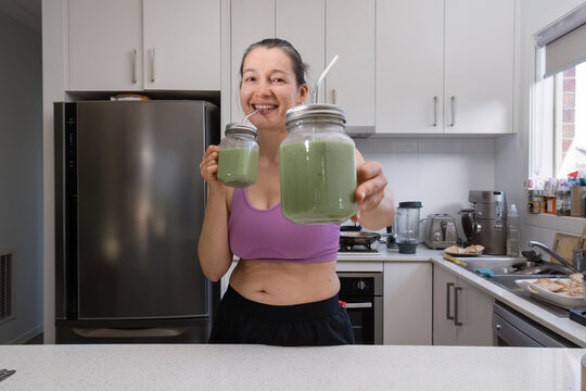 Happy Unfit Woman In Sports Clothing Drinking Green Smoothie And Offering One Glass Of Smoothie To You.