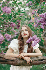 Obraz na płótnie Canvas Young pretty girl in white dress with long hair standing among blooming branches of lilac in spring garden or park looking up, outdoor lifestyle romantic portrait, idea of freshness, relax and resting
