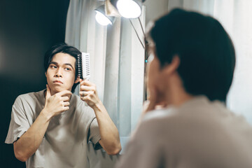 Young adult asian man using comb for hairstyle in this mirror reflection at home.