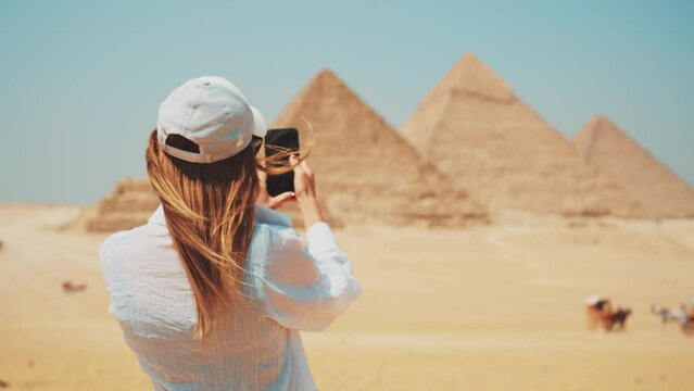 Tourist woman in takes photo on smartphone of Great Giza pyramids in Cairo Egypt enjoys attraction of world UNESCO ancient heritage, back view. Tourism travel wanderlust journey sightseeing concept.