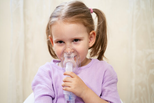 Cute little girl inhaling mist form nebulizer at home. Holding mask over her nose and mouth 