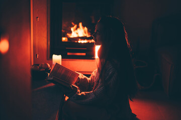 Cozy home. Young woman reading book near the fireplace.