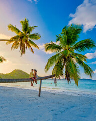 Praslin Seychelles tropical island with white beaches and palm trees, a couple of men and woman watching sunset above a palm tree climbing in a palm tree at Anse Volber Seychelles. - obrazy, fototapety, plakaty