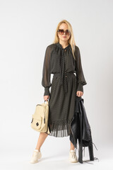 A woman in sunglasses and a casual dress holds a jacket and backpack in her hands