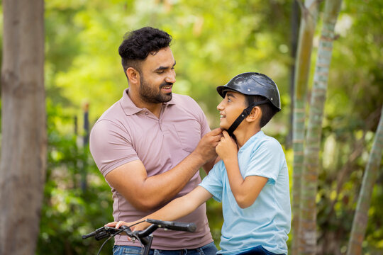 Happy indian father puts protection helmet on son for bicycle ride at park - concept of caring father, Parental guidance and Bicycle safety