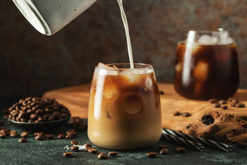 Ice coffee in a tall glass with cream poured over, ice cubes and beans on a dark concrete table.