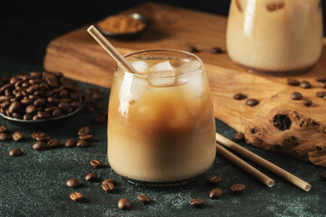 Ice coffee in a tall glass with cream poured over, ice cubes and beans on a dark concrete table.