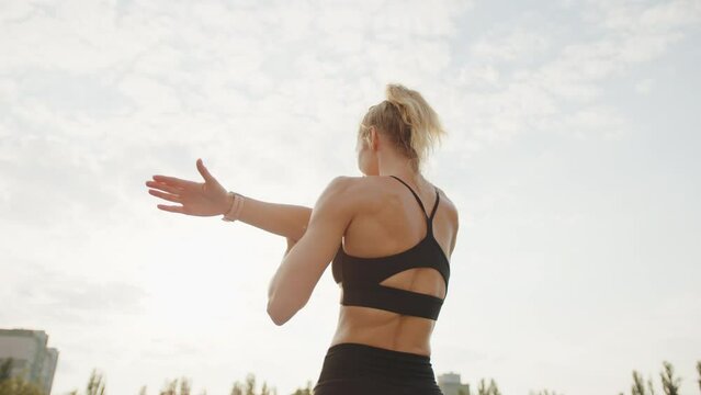 Low angle, back view of sporty blonde performing arms stretching exercises in the stadium. Clear sky, tops of trees, skyscraper in the background. High quality 4k footage