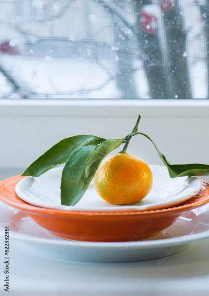 Poster tangerine with a branch and leaves on a plate against the background of a winter window with the sno