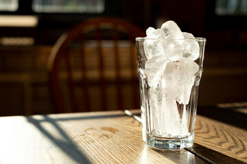 Ice cubes in clear glass placed on wooden table in kitchen, natural sunlight shines from behind.
