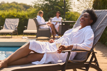 Mature Woman Wearing Robe Outdoors On Lounger Drinking Champagne On Spa Day
