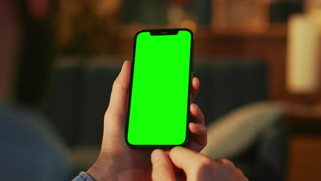 Man's hands holding smartphone with green screen on blurred background of living room in evening. Isolated mobile phone. Man touching cellphone, scrolling with finger on screen of gadget. Indoors