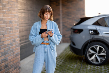 Young woman using smart phone while charging her electric car near garage of her house. Concept of...
