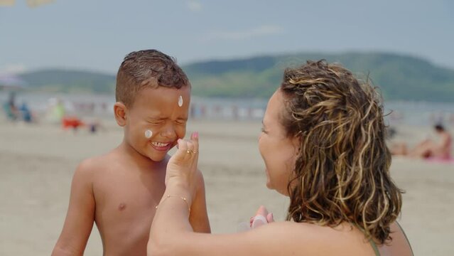 mother applying sunblock lotion on sons skin for sun protection little boy getting ready to swim on the beach with mom using sunscreen caring for childs health on sunny day 
