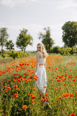 Blonde young woman standing in a field with wild red poppies, wearing a white dress, smiling, looking at camera.