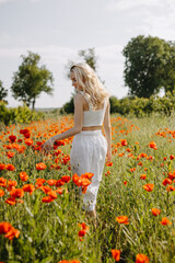 Blonde young woman standing in a field with wild red poppies, wearing a white dress.