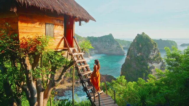 Silhouette beautiful travel girl in dress rising to a picturesque tree hut on top mountain cliff on background tropical beach and scenery rocks in ocean 4K