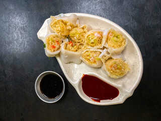 Top view of steamed vegetable and shrimp springrolls on a plate, on textured black table in restaurant. Dish is accompanied by black sweet sauce and chilli sauce