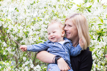 young smiling mother holding her cute cheerful son and enjoying blooming trees in the park at spring