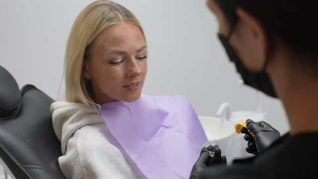 Close-up view from shoulder of unrecognizable female orthodontist doctor showing aligners or kappa for teeth alignment to young woman patient, sitting on dental chair in dentistry clinic, slow motion.