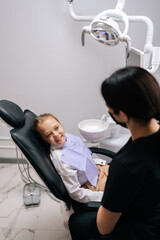 Vertical shot of unrecognizable female dentist talking with smiling child girl lying on dental chair in front of doctor, looking with happy expression. Concept of children health care treatment.