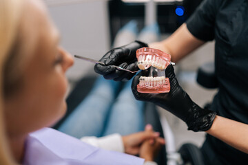 Close-up cropped shot of unrecognizable orthodontist in rubber gloves showing artificial human jaws with dental braces for teeth correction to female patient, visiting doctor orthodontic issues.