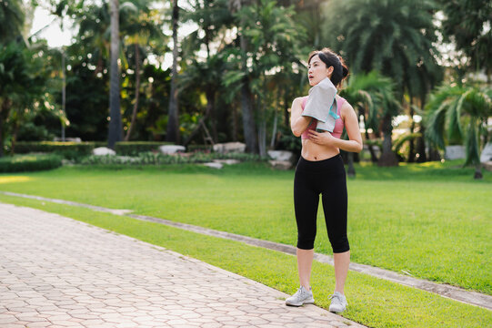 Female jogger. Get inspired fit active image of 30s Asian young woman in pink sportswear use small towel in park after run, enjoying healthy outdoor activity. Wellness and fitness in natural setting.