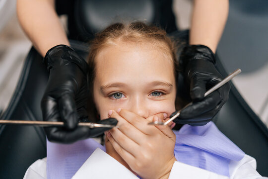 Close-up portrait of little cute child girl closing mouth by fear in dental clinic, frightened by dental drill, afraid of treatment. Cropped shot of unrecognizable female doctor holding dental tool.