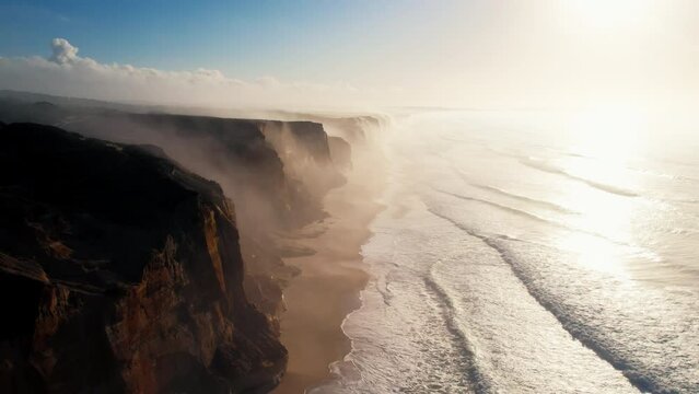 Early sunrise aerial drone shot of misty and sunny empty sand beach. Epic and cinematic high drone perspective of waves crushing and rolling over wild secluded beach