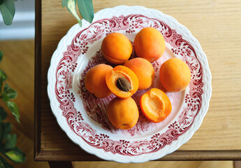 Fresh ripe apricots on plate on wooden table, sunny morning, village atmosphere, rustic. Cosy home interior. Harvest concept. Pile of whole juicy orange apricots and one halved apricot on table